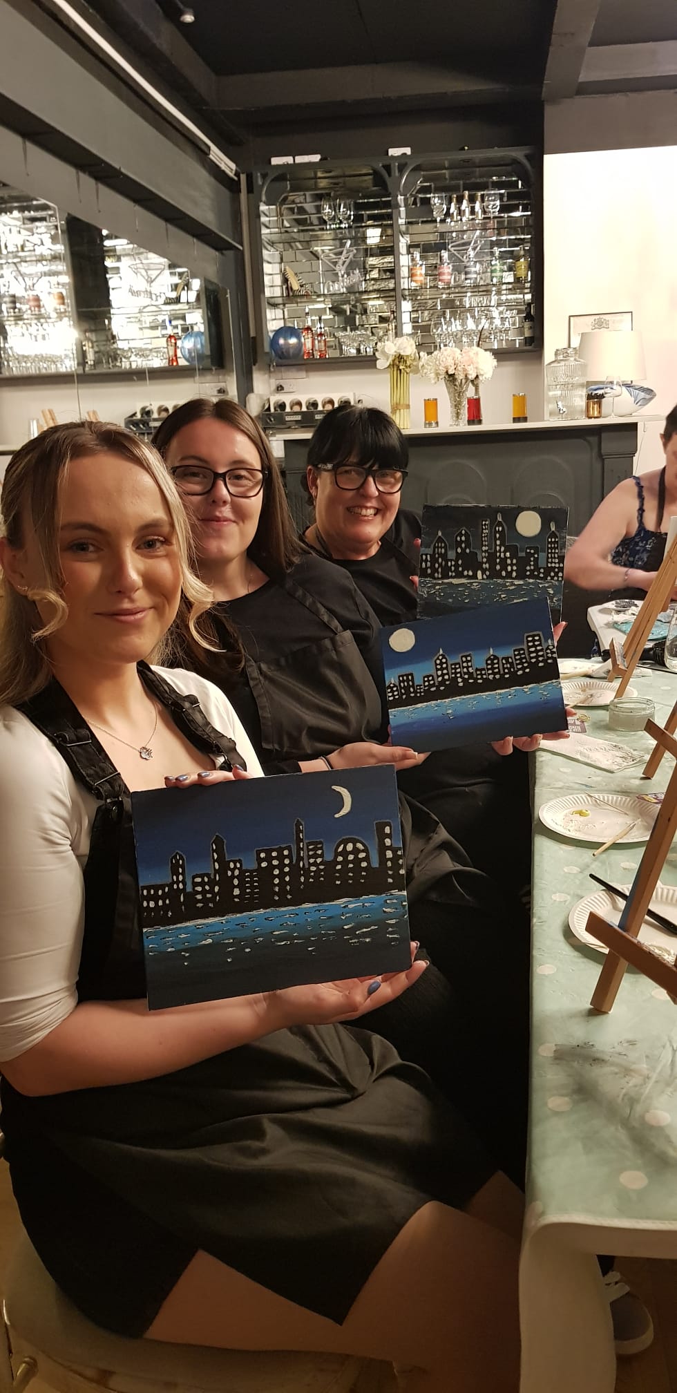 Three people at a Paint and Sip session at Satchmo's Corner in Rugby smiling and proudly holding up their 'evening cityscape' paintings.