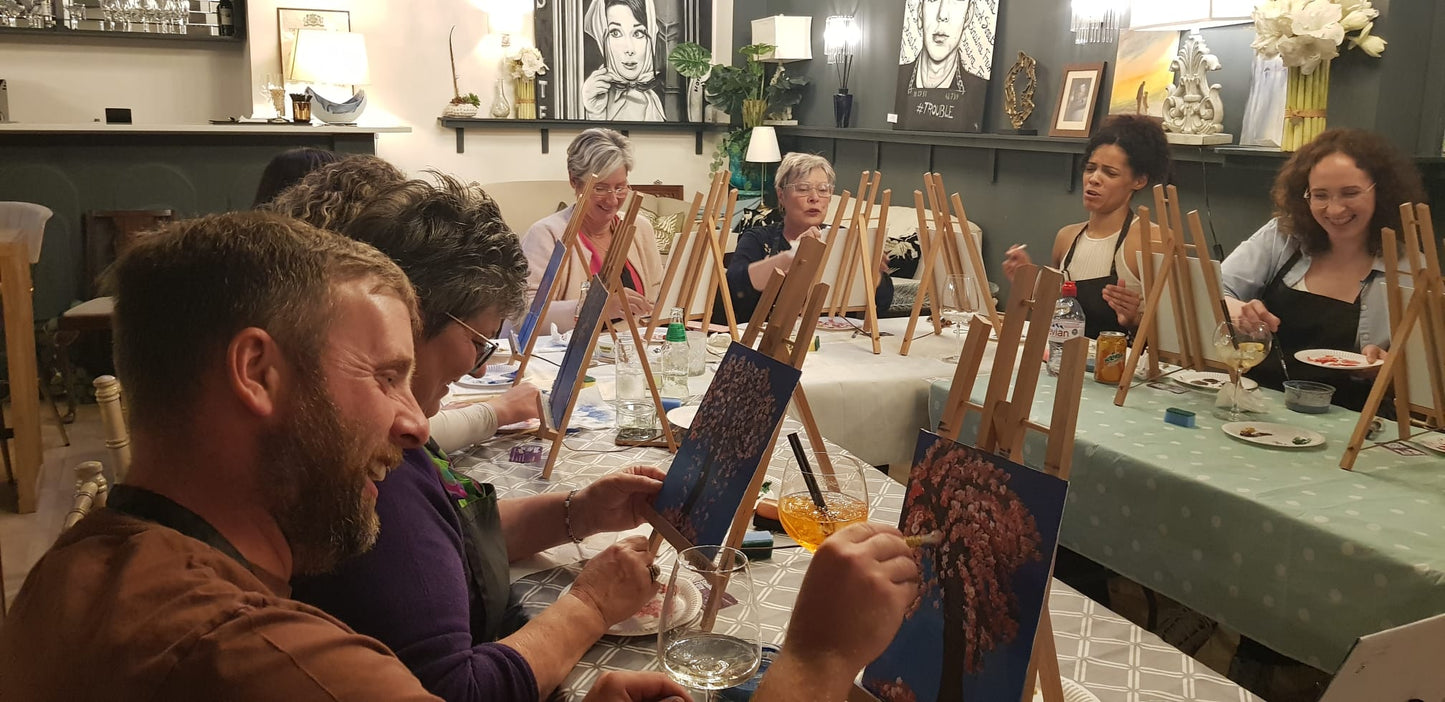 Group of people smiling and laughing at a Paint & Sip session in Rugby while painting their own pink blossom trees at individual easels.