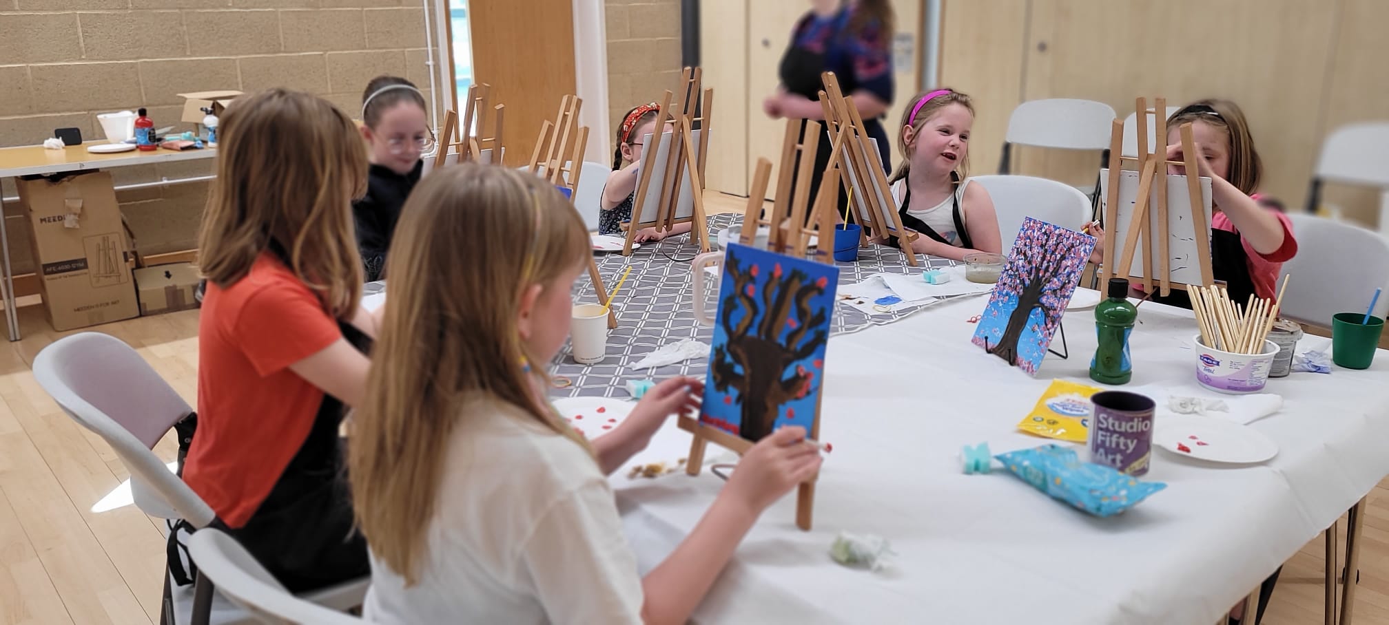 Six children sat at a table engaged in painting activities at a Junior Paint & Sip session run by Studio Fifty Art