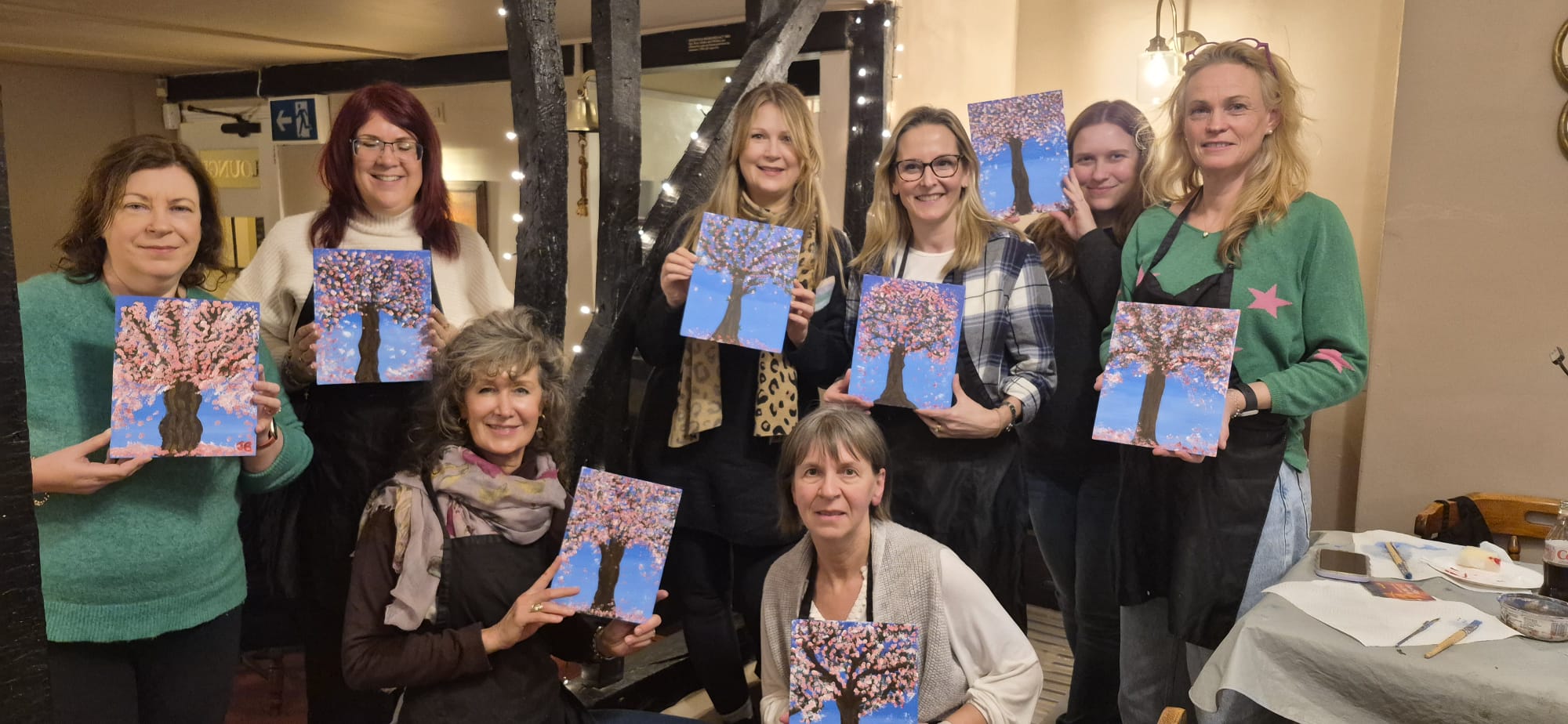 Group of women smiling and holding up their paintings of Blossom Trees, created at a Paint & Sip event at a cosy local pub with twinkling lights in the background