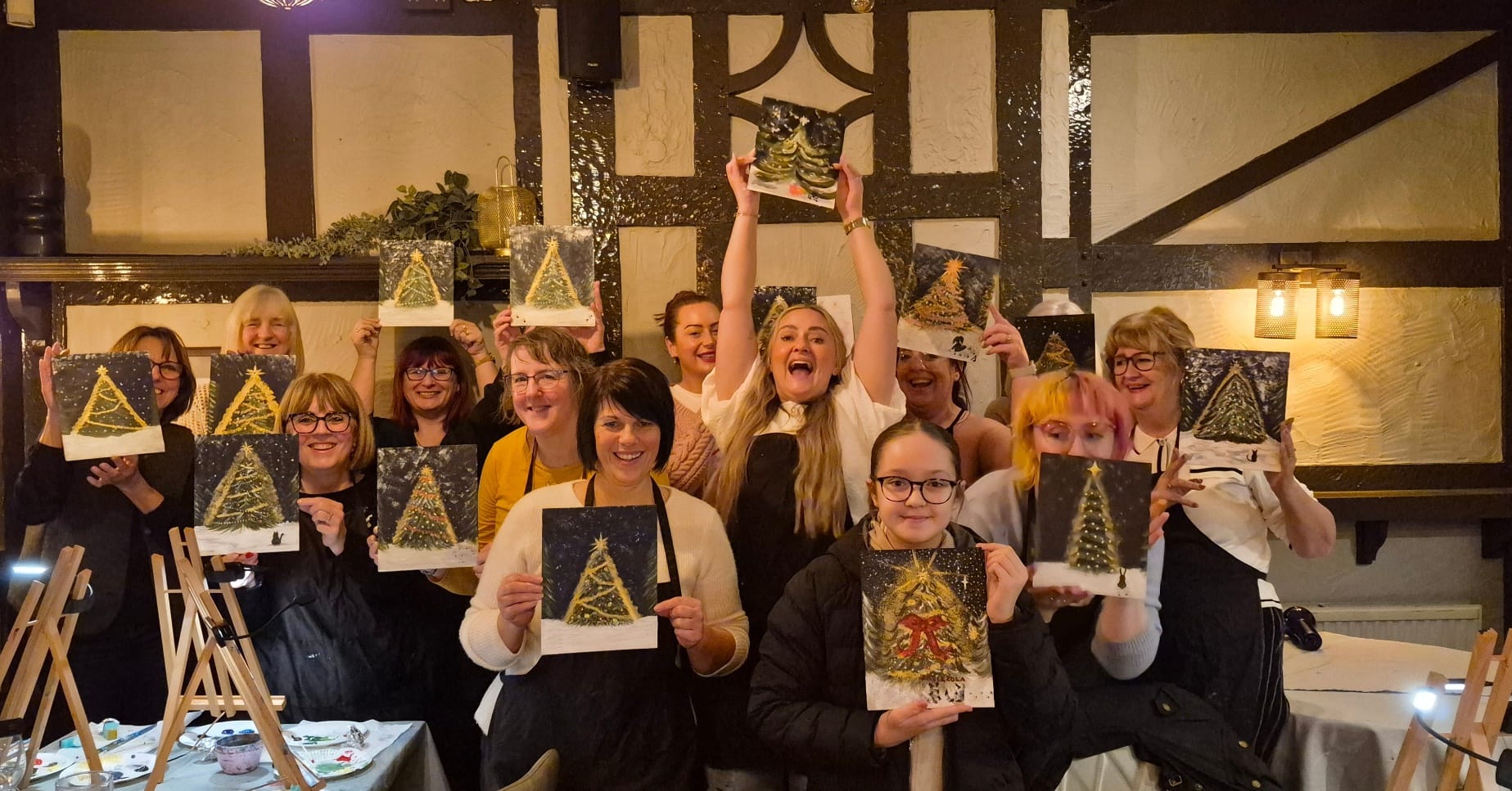 Group of 12 people standing together in a cosy pub setting proudly holding up their paintings of Christmas trees while smiling and laughing together