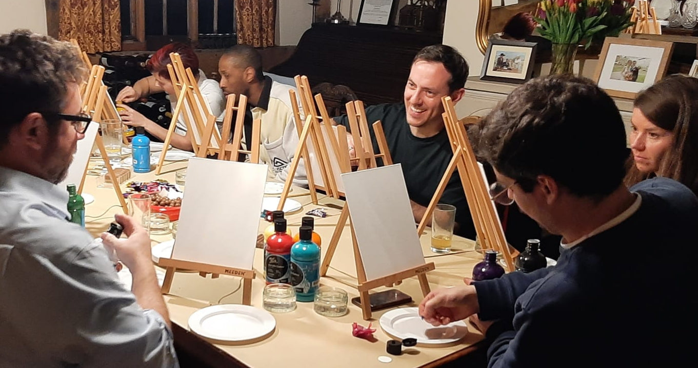 Group of people sat at a table with easels and canvases, laughing and smiling and about to start painting at a Paint & Sip event