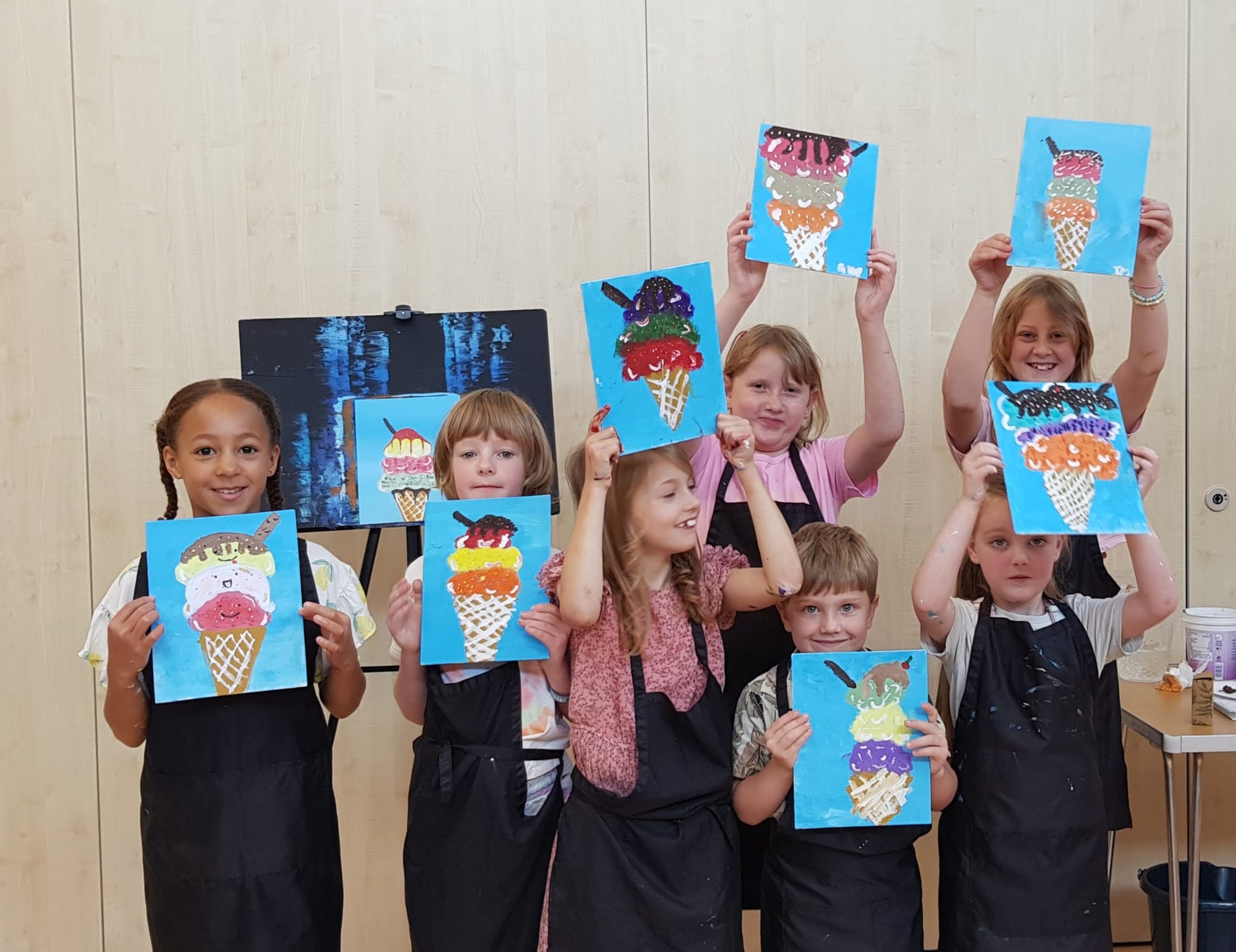 Group of seven children at a Studio Fifty Art Junior Paint & Sip session smiling, laughing and proudly holding up their paintings of ice creams.