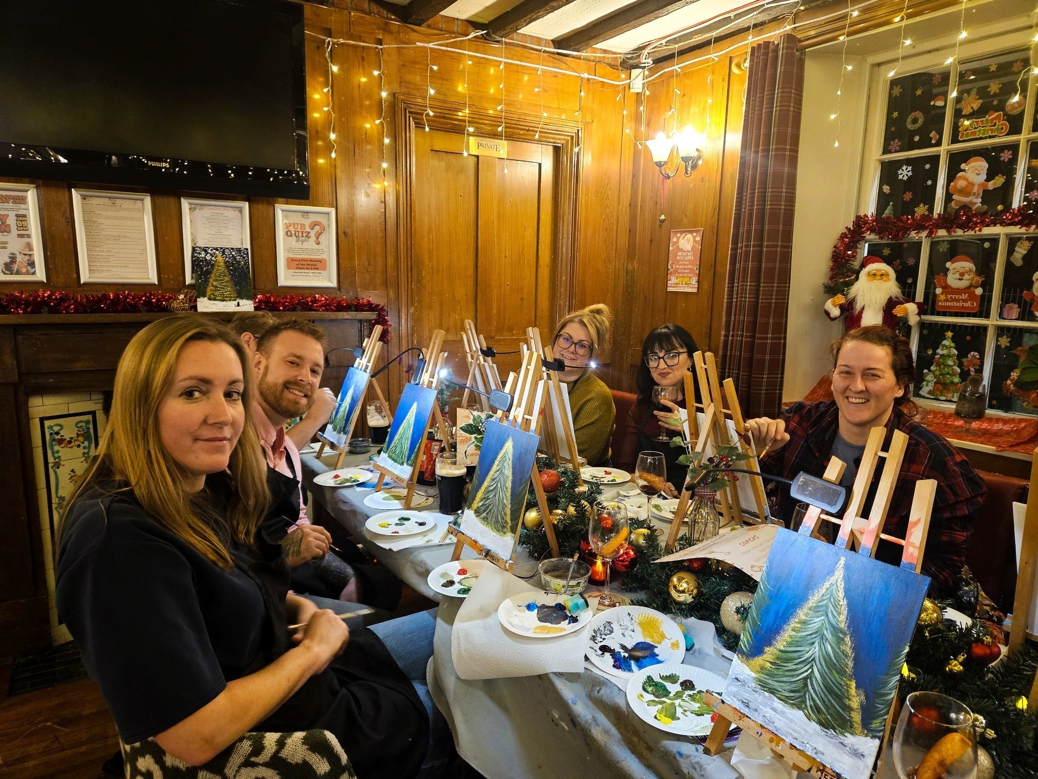 Group of people sitting around a table  in front of easels and painting a Christmas tree at a Paint & Sip session in a cozy Norwich pub with Christmas decorations.