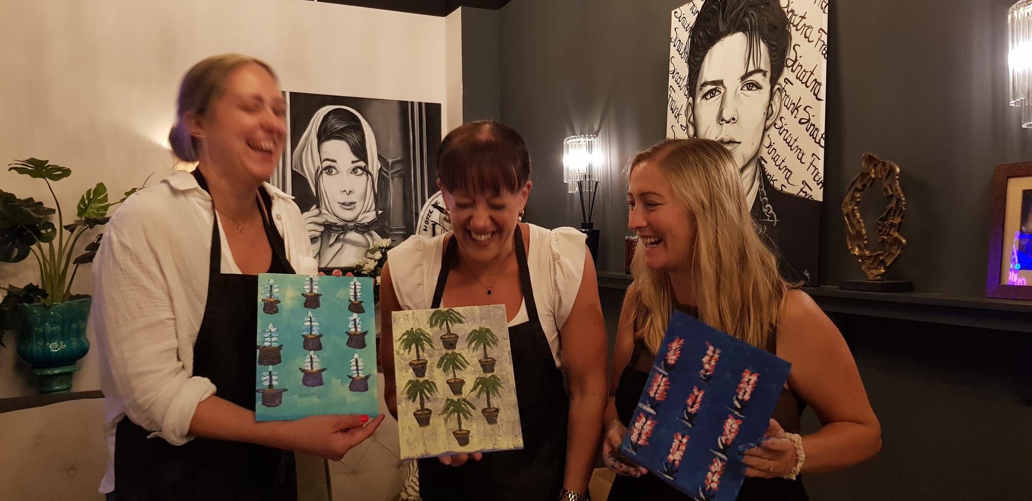 Three women  laughing together and holding their artwork of tree patterns up.  They are stood in a room with artistic decor after a Paint & Sip night.