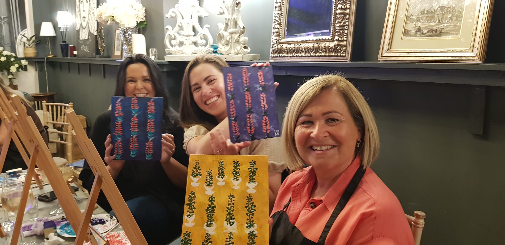 group of 3 women at a Paint & Sip event smiling and laughing while proudly holding up their paintings of tree patterns 