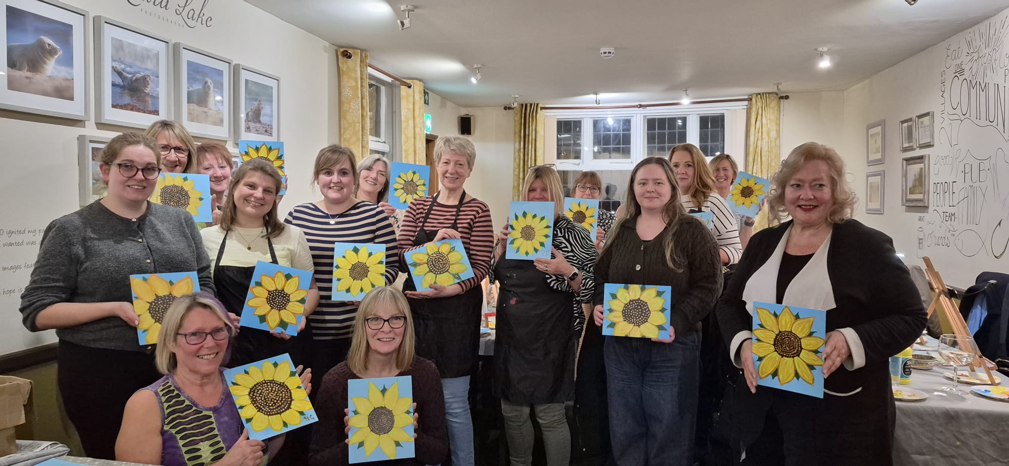 Large group of people in a pub setting, smiling and proudly holding up canvases of their own painted sunflowers they have just completed during a Paint & Sip session at The White Horse in Chedgrave