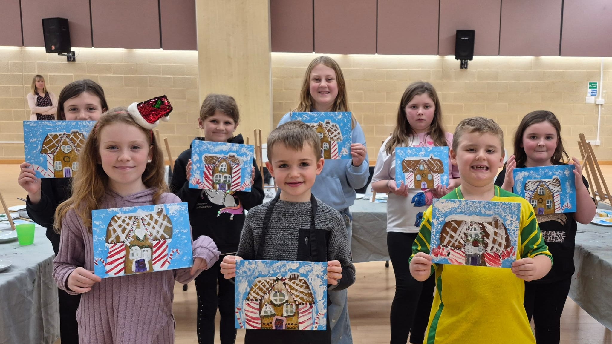 Large group of children at a holiday art class, smiling and proudly holding up their paintings of a gingerbread house.