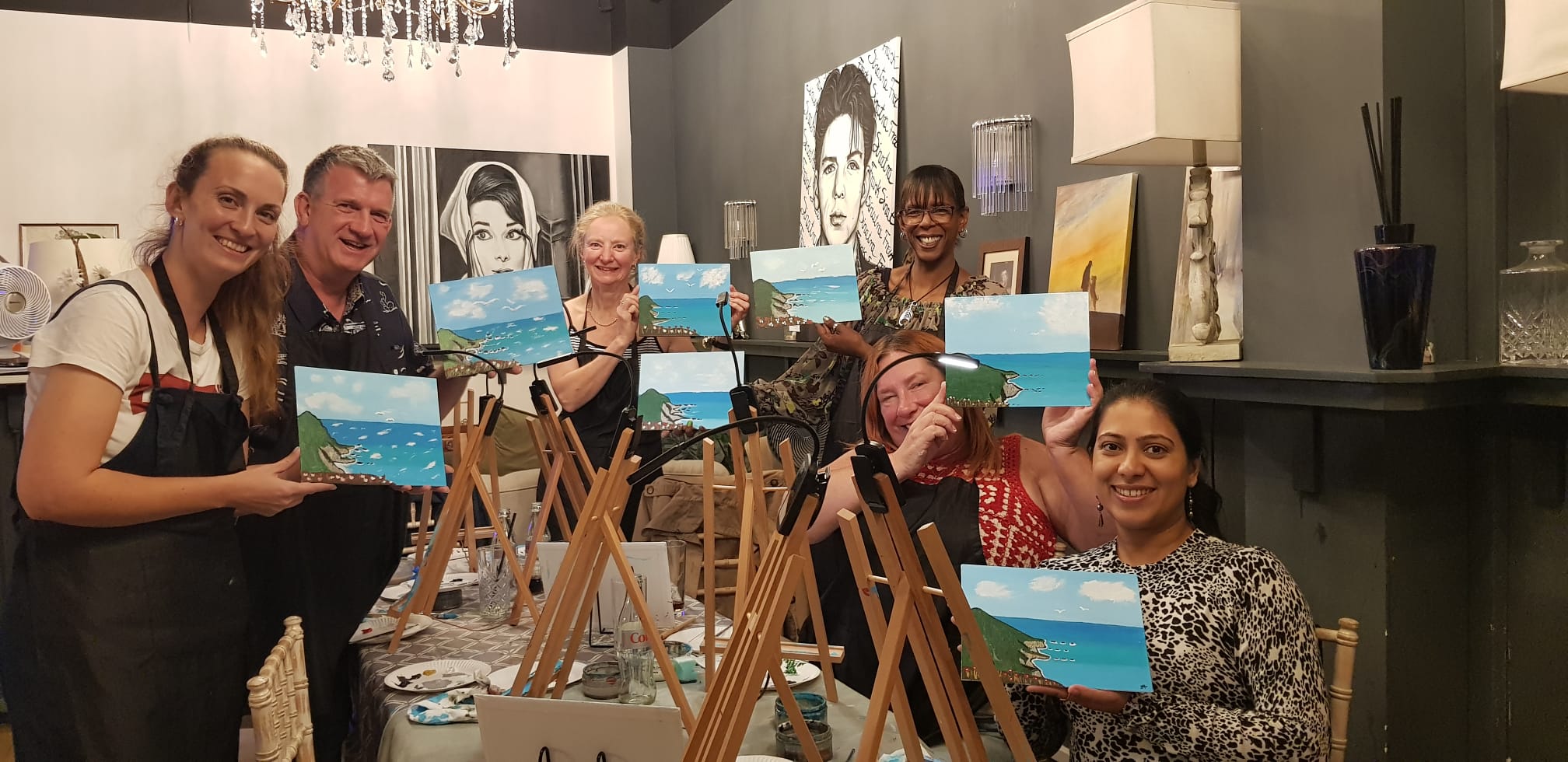 Group of 6 people standing in an art filled restaurant setting at a table with easels on it smiling and laughing, holding up their paintings of seascapes  they've just completed at a Paint & Sip night