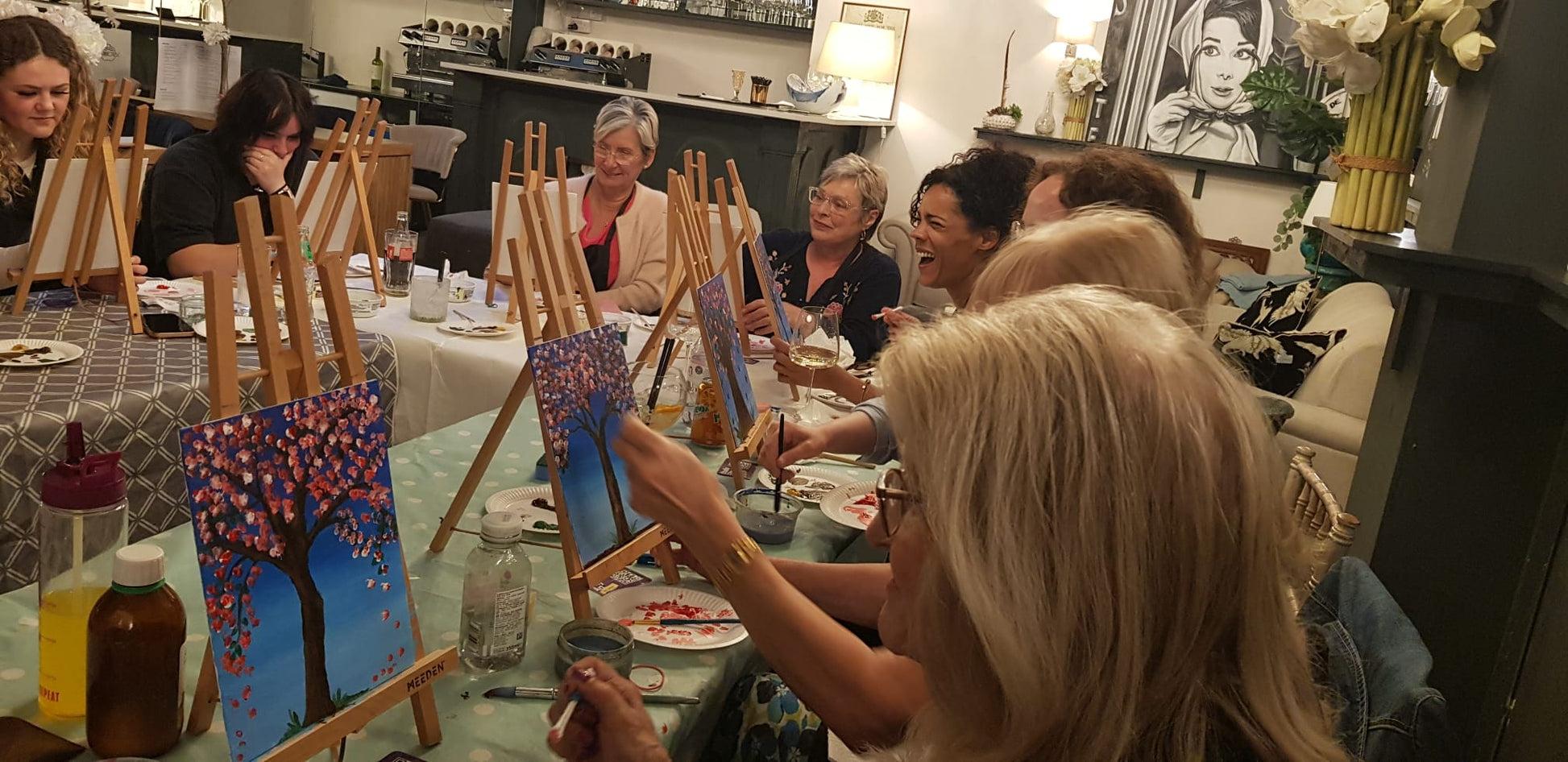 Group of people smiling and laughing sat round a table at easels, painting blossom trees on canvases at a Paint & Sip session